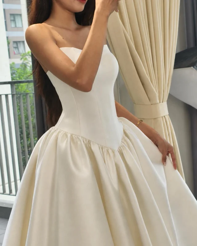 Điều gì làm nên chi phí của một chiếc váy cưới may đo Cô dâu mặc váy cưới satin ivory thân trên ôm sát hạ eo, chân váy xoè phồng, chụp bên rèm cửa studio