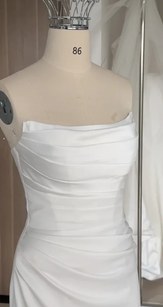 Thân váy cưới satin trắng xếp ly chéo, cổ ngang trễ vai, đang hoàn thiện trên ma-nơ-canh
