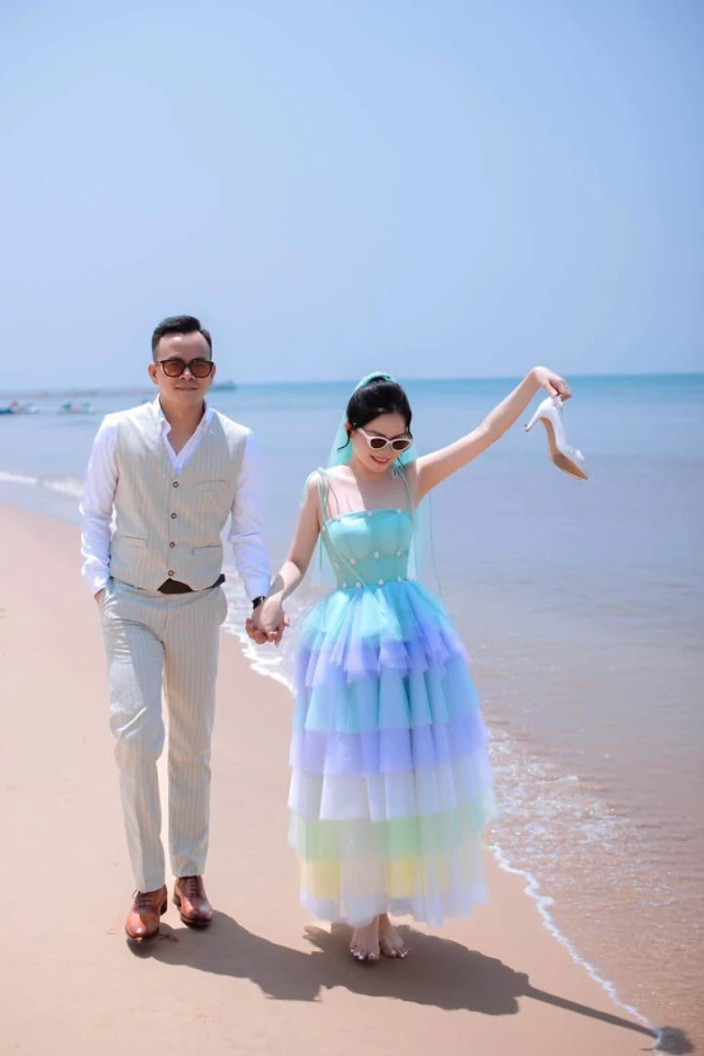Váy cưới nhiều tầng voan lưới phối tím và xanh ngọc Dâu cầm giày đi chân trần trên bãi biển, các tầng voan tím, xanh ngọc nổi bật dưới nắng