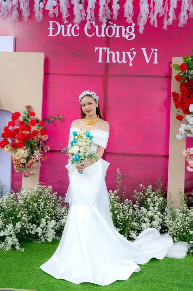 Dâu cầm hoa trước phông hồng, đuôi váy satin trải dài trên thảm cỏ